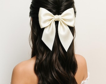 May include: A large, ivory satin bow hair clip. The bow is centered on dark brown wavy hair. The hair clip is a simple, elegant accessory. The bow's smooth texture and color suggest a formal or bridal style.