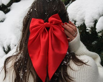 May include: A large, vibrant red bow is the focal point, set against a backdrop of snow-covered evergreen branches. The bow is attached to the back of the person's head, with dark hair and a white sweater visible. The bow's bold color contrasts with the winter scene.