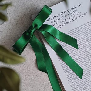 May include: A vibrant emerald green satin ribbon bow, elegantly tied and resting on a book. The book's pages display printed text. The bow's glossy texture and rich color create a striking contrast.