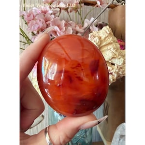 May include: A polished, oval-shaped carnelian crystal sphere. The stone is a vibrant orange-red color with darker, reddish-brown inclusions. The surface is smooth and reflective, showcasing the natural patterns within the crystal. The text "Violet Veil Creations LLC" is visible.