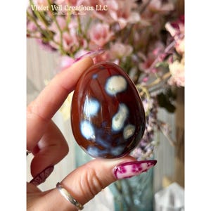 May include: A polished, egg-shaped stone with a deep burgundy base and white and blue patterns. The stone is held in a hand, with the text "Violet Veil Creations LLC" visible in the background. Crystals & Spellwork.