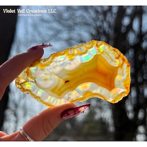 May include: A translucent, polished agate slice with vibrant orange and white bands. The stone is held up to the light, revealing iridescent rainbow hues. The text "Violet Veil Creations LLC CRYSTALS & SPELLWORK" is visible in the upper left corner.
