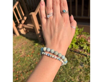 May include: Two beaded bracelets with blue and white stones are stacked on a wrist. The image also shows several rings on the fingers. The text "Violet Veil Creations" is at the bottom of the image.