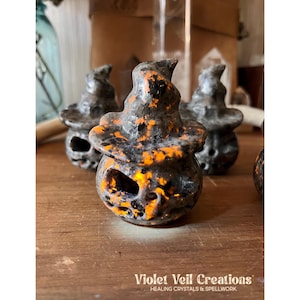 May include: Close-up of several carved stone pumpkins wearing witch hats. The pumpkins are black with orange and yellow accents. The text "Violet Veil Creations" is at the bottom of the image.