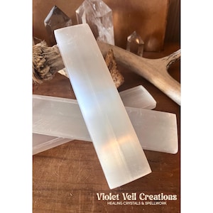May include: Several rectangular, translucent white selenite crystal wands are displayed on a wooden surface. The wands are the focal point, with other crystals and natural elements in the background. The image includes the text "Violet Veil Creations, Healing Crystals & Spellwork."
