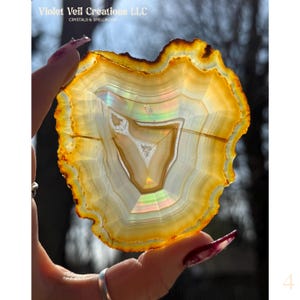 May include: A translucent, polished agate slice with concentric bands of yellow, orange, and white. The stone has a natural, irregular shape and displays a central, triangular pattern. The text "Violet Veil Creations LLC" is visible.