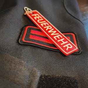 Könnte beinhalten: Nahaufnahme einer dunkelblauen Marinejacke mit einem rot-schwarzen Aufnäher und einem roten Schlüsselanhänger. Der Schlüsselanhänger und der Aufnäher tragen beide das Wort "FEUERWEHR" in weißen Buchstaben. Die Jacke hat einen Klettverschluss.
