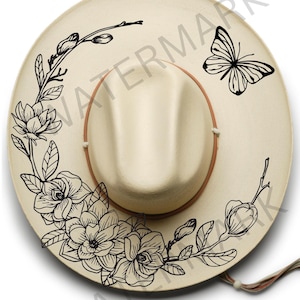 May include: A beige cowboy hat with a black floral design and a butterfly on the side. The design includes magnolia flowers and leaves.