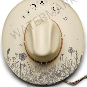 May include: Cream-colored fedora hat with a brown brim and a decorative floral and celestial design. The hat features a detailed illustration of wildflowers and celestial bodies, including moons and suns. The hat has a decorative band and a chin strap.