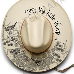 May include: A cream-colored hat with a brown brim and the words "enjoy the little things" in black script. The hat is decorated with black and white floral and bird designs. A decorative hat for everyday wear.