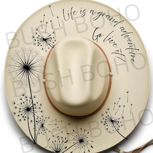 May include: Cream-colored fedora hat with a brown band and a rounded crown. The hat features black dandelion illustrations and the text "Life is a grand adventure. Go live it!" in a script font. A stylish accessory.