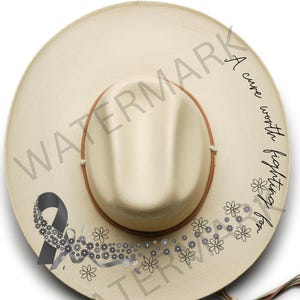 May include: A cream-colored hat with a brown band and brim. The hat features a black ribbon and floral design, along with the text "A cure worth fighting for". The hat is decorated with small flowers and rhinestones.