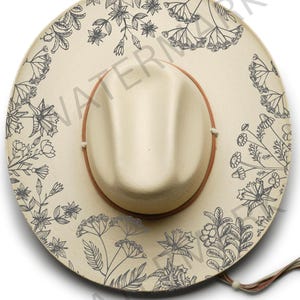 Puede incluir: Un sombrero beige de ala ancha con un diseño floral negro. El sombrero tiene una banda de cuero marrón y una barbilla de cuero marrón.