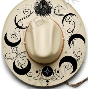 May include: Cream-colored cowboy hat with a decorative celestial design. The hat features black crescent moons, stars, and a woman's silhouette. A brown band encircles the crown, and a thin strap is attached. The hat is from Bush Boho Hat Burning Stencils.