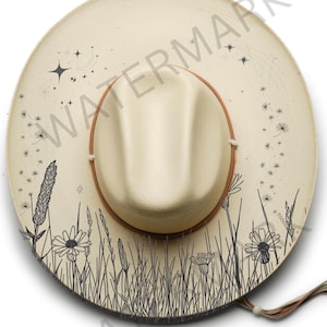 May include: Cream-colored hat with a brown brim and a beige crown. The hat is decorated with black line drawings of wildflowers, grass, stars, and dandelions. The hat has a string tie and a logo that says "BUSH BOHO HAT BURNING STENCILS."