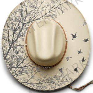 May include: A cream-coloured fedora hat with a brown leather band and a decorative design. The hat features a tree branch and bird pattern in grey. The hat has a brim and a beige crown.