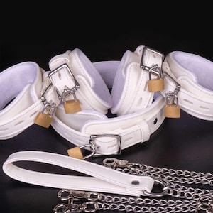 Op de afbeelding: Witte leren boeien met zilveren gespen en hangslotjes. De boeien zijn verbonden door een witte leren band met een zilveren gesp. Een witte leren riem met een zilveren ketting is inbegrepen.