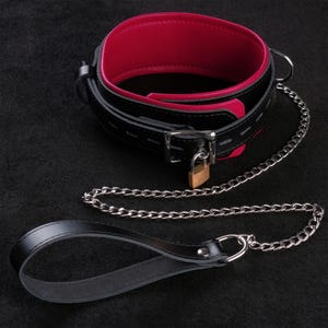 May include: A black and red leather collar with a silver chain leash. The collar has a buckle closure and a small padlock. The interior of the collar is red. The leash has a loop handle.