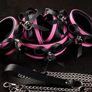 Peut inclure: Ensemble de bondage noir et rose comprenant des entraves pour les poignets et les chevilles, un collier et une laisse avec une chaîne argentée. L'ensemble est orné de nœuds noirs et d'anneaux en métal argenté.