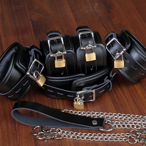 Op de afbeelding: Een set zwarte leren handboeien met zilveren sloten en een bijpassende zwarte leren riem met een zilveren ketting.