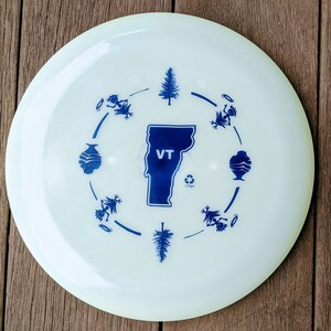 May include: White frisbee with a blue outline of the state of Vermont and the letters "VT" inside. The frisbee has a blue circle around the state with blue images of trees, mountains, and people.