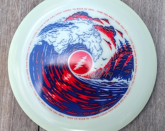 May include: A pale green disc featuring a graphic of a large blue and red ocean wave crashing near a forest. A red circle with a white lightning bolt is in the centre. The text reads: LISTEN TO THE RIVER SING SWEET SONGS, TO ROCK MY SOUL, FARE YOU WELL, FARE YOU WELL. I LOVE YOU MORE THAN WORDS CAN TELL. RIP ROBERT HUNTER.