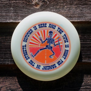 May include: A white frisbee with a blue and orange design. The design features a skeleton dancing on a road with a sun and the text "THE TIME IS RIGHT FOR DANCIN' IN THE STREETS. SUMMER IS HERE AND"