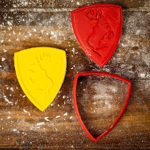 May include: Two red and one yellow plastic cookie cutters shaped like shields with a moose design inside. The cutters are on a brown wooden surface covered in white flour.