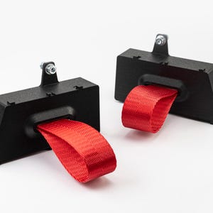 May include: Two black metal brackets with red fabric loops. Each bracket has a mounting tab with a bolt. The brackets are angled and have a rectangular opening for the red fabric loop.