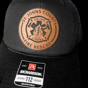 May include: Black trucker hat with a brown leather patch featuring the St. Johns County Fire Rescue logo. The logo includes a cross with an axe and a hose, the words "St. Johns County", "Est. 1998", "Florida", and "Fire Rescue". The hat also has a black adjustable strap with the Richardson logo and the text "Est. 1970", "Richardson", "Original 112 Trucker", and "Adjustable".