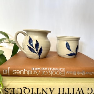 May include: A cream-colored ceramic pitcher and small cup set. Both pieces feature a hand-painted blue leaf design. The pitcher has a handle and a spout. The set is displayed on a vintage book.
