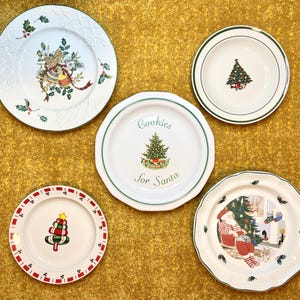 May include: A collection of five white ceramic Christmas plates. The plates feature festive designs including a Christmas tree, Santa Claus, and holly. One plate has the text "Cookies for Santa". Each plate has a green or red border.