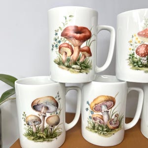 Pu&ograve; includere: Tazze in ceramica bianca con illustrazioni di funghi. Le tazze presentano vari disegni di funghi nelle tonalit&agrave; del marrone, del rosso e del giallo, con erba verde e fiori blu. Le tazze sono impilate.