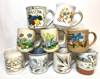 May include: A collection of nine ceramic mugs stacked in a pyramid arrangement. The mugs feature various designs including the word GRANDPA with fruit, floral patterns, a house in a winter landscape, seashells, wheat stalks, and a cow in a field.