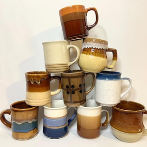 May include: A collection of ten ceramic mugs stacked in a pyramid formation. The mugs feature various earth-toned glazes, including shades of brown, tan, cream, and blue, with patterns such as horizontal stripes, speckles, and geometric shapes.