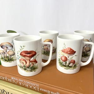 Pu&ograve; includere: Cinque tazze in ceramica bianca con illustrazioni di funghi. Ogni tazza presenta un diverso design di funghi nelle tonalit&agrave; del marrone, del rosso e del beige, con fogliame verde. Le tazze hanno un design classico con manico e sono esposte su una pila di libri.