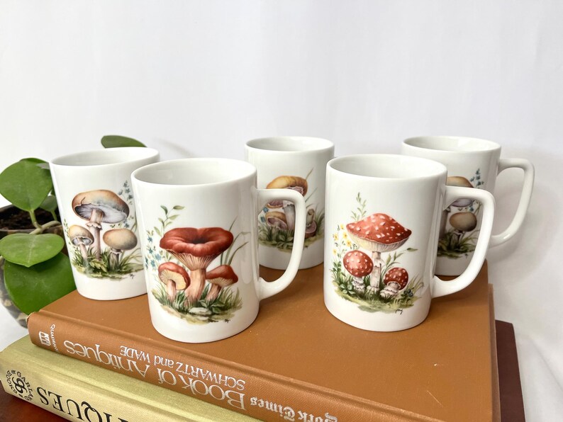 Pu&ograve; includere: Cinque tazze in ceramica bianca con illustrazioni di funghi. Ogni tazza presenta un diverso design di funghi nelle tonalit&agrave; del marrone, del rosso e del beige, con fogliame verde. Le tazze hanno un design classico con manico e sono esposte su una pila di libri.