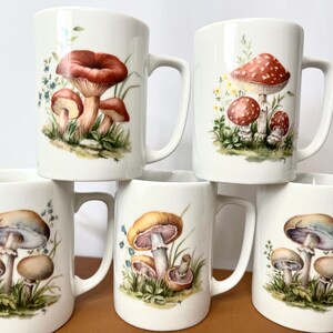 Pu&ograve; includere: Una collezione di sei tazze in ceramica bianca, ognuna decorata con un'illustrazione ad acquerello di funghi diversi. I disegni dei funghi presentano vari colori, tra cui rosso, marrone e beige, su uno sfondo di erba verde e piccoli fiori blu.