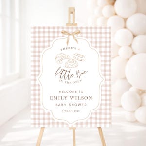 May include: A baby shower welcome sign on an easel. The sign has a tan and white gingham background with a white, ornate frame. The text reads "There's a Little Bun in the Oven" and welcomes Emily Wilson to her baby shower on April 5th, 2026.