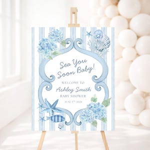 May include: A baby shower welcome sign on a wooden easel. The sign has a blue and white striped background with watercolour illustrations of seashells, starfish, and hydrangeas. Text reads "Sea You Soon Baby!" and baby shower details.