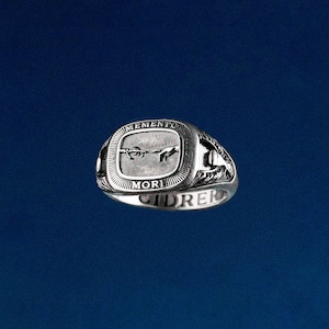 May include: A silver ring with a rectangular face featuring a depiction of two hands reaching towards each other, with the words "MEMENTO MORI" inscribed. The ring has a detailed design on the sides and the word "CIDRER" on the band.