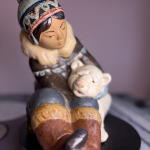 May include: A ceramic figurine of a person in winter attire, seated and embracing a polar bear cub. The figure wears a patterned hat, a fur-trimmed coat, and brown boots. The polar bear is white with a blue scarf.