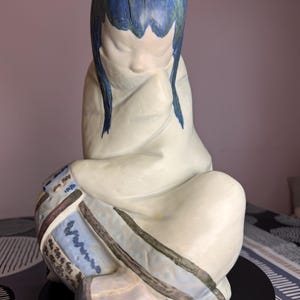 Puede incluir: Figura de cerámica de una figura sentada con cabello azul y una túnica blanca. La figura está en una pose contemplativa, con la cabeza inclinada y las manos juntas. La túnica tiene un diseño estampado en azul, marrón y blanco.