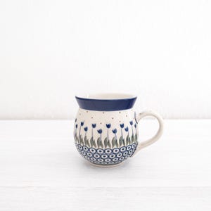 May include: A white ceramic mug with a blue rim and a floral pattern. The pattern features blue tulips with green leaves and white dots.