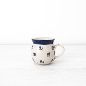 May include: A white ceramic mug with a blue rim and a blue and green floral pattern. The mug has a rounded body and a handle.