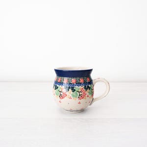 May include: A white ceramic mug with a blue rim and a floral pattern in shades of blue, green, pink, and yellow. The mug has a rounded body and a handle.