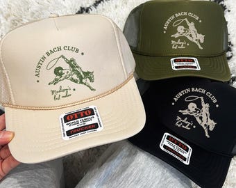 May include: Four trucker hats in beige, olive green, and black, with mesh backs and curved brims. Each hat displays the text "AUSTIN BACH CLUB" and a rodeo graphic. The hats have "OTTO" labels.