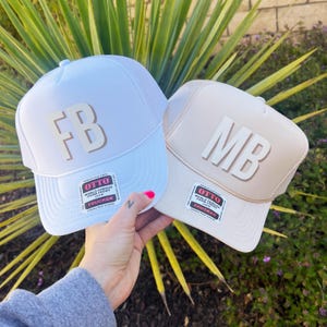 May include: Two trucker hats, one white and one beige, are held up against a background of green plants. The white hat has "FB" in tan letters, and the beige hat has "MB" in white letters. Both hats have a mesh back and a "OTTO" label.
