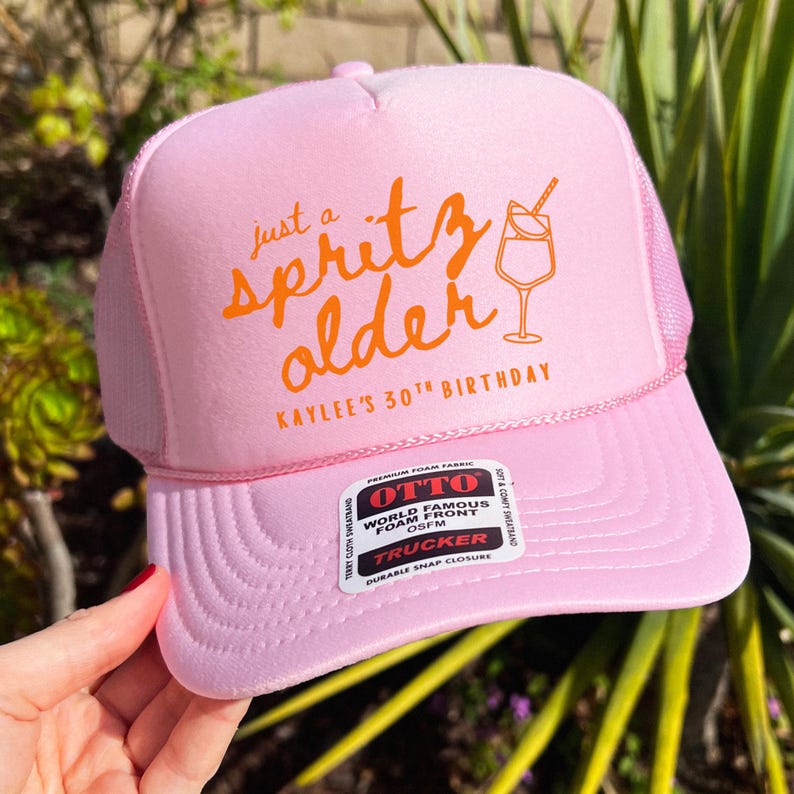 May include: Pink trucker hat with the text "just a spritz older" and a cocktail illustration in orange. The hat also includes the text "Kaylee's 30th Birthday". The hat has a mesh back and a snap closure.
