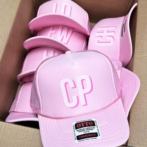 May include: A collection of pink trucker hats with white lettering and mesh backing. The hats feature the letters "CP", "FW", "CH", and "ID". Each hat has a curved brim and a snap closure. The hats are stacked inside a cardboard box.